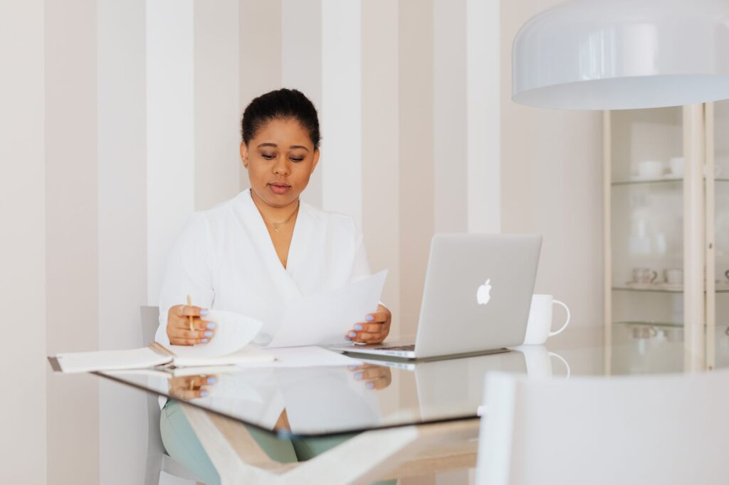 Mujer de negocios en una oficina moderna, revisando documentos en un escritorio de cristal con un portátil.